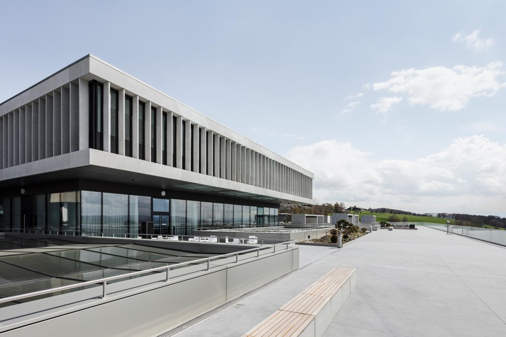 Ein modernes Geb&auml;ude mit gro&szlig;en vertikalen Fenstern und Betonelementen steht auf einer gro&szlig;z&uuml;gigen Terrasse mit Glasgel&auml;nder und B&auml;nken, die den Blick auf gr&uuml;ne Felder freigibt - ein idealer Ort zum G&auml;rtnern oder f&uuml;r eine ruhige Therapie bei teilweise bew&ouml;lktem Himmel.