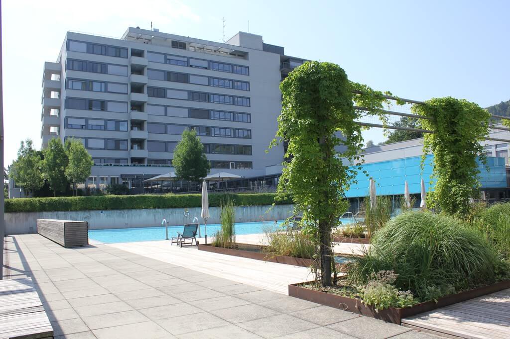 Ein modernes Geb&auml;ude mit mehreren Etagen steht hinter einem Au&szlig;enschwimmbad, umgeben von Gr&uuml;nfl&auml;chen, die ideal zum G&auml;rtnern sind, Liegest&uuml;hlen und schattigen Bereichen, die sich an einem sonnigen Tag perfekt f&uuml;r eine Therapie eignen.