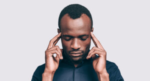 Ein Mann mit kurzen Haaren und gestutztem Bart dr&uuml;ckt seine Zeigefinger an die Schl&auml;fen, die Augen in tiefer Konzentration geschlossen, als versuche er, seine Gehirnleistung zu steigern, vor einem schlichten hellgrauen Hintergrund.