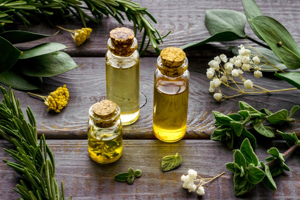 Drei kleine Glasflaschen mit Korkstopfen, gef&uuml;llt mit gelben und klaren &Ouml;len, umgeben von frischen gr&uuml;nen Kr&auml;utern und kleinen wei&szlig;en und gelben Blumen auf einer rustikalen Holzoberfl&auml;che - perfekt f&uuml;r Aromatherapie und die Heilkraft wohltuender D&uuml;fte.