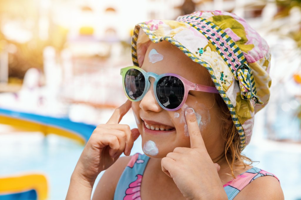 Ein l&auml;chelndes Kind, das eine Sonnenbrille und einen gebl&uuml;mten Hut tr&auml;gt, tr&auml;gt Sonnencreme auf seine Wangen auf, w&auml;hrend es an einem sonnigen Sonnenscheintag in der N&auml;he eines Pools steht - um seine Haut zu sch&uuml;tzen und Nebenwirkungen zu vermeiden.