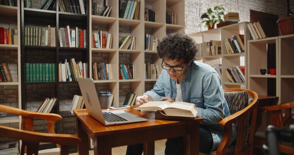 Ein junger Mann mit lockigem Haar und Brille studiert an einem Holztisch mit einem aufgeschlagenen Buch, einem Notizbuch und einem Laptop in einer mit B&uuml;cherregalen gef&uuml;llten Bibliothek. Eine Kaffeetasse, eine Topfpflanze und ein Nootropika zur Steigerung der Gehirnleistung stehen ebenfalls auf dem Tisch.
