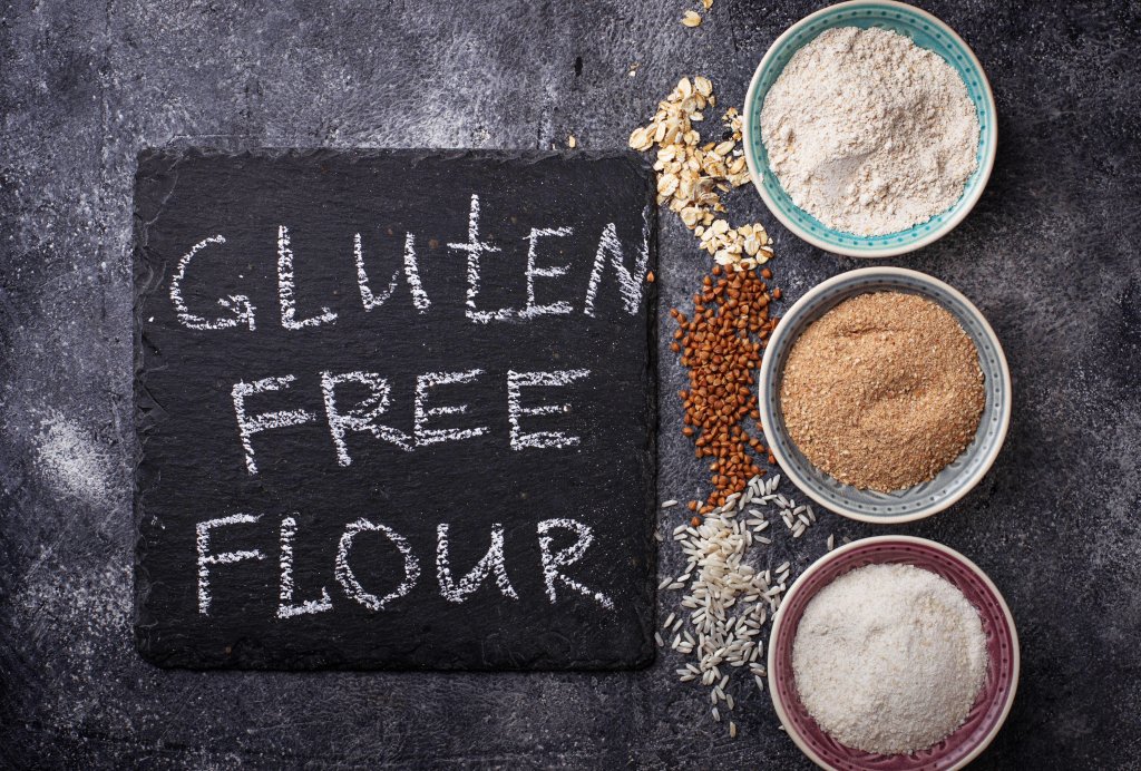 Drei Sch&auml;lchen mit verschiedenen glutenfreien Mehlen, umgeben von Reis, Hafer und Samen, sind neben einer schwarzen Tafel mit der Aufschrift GLUTEN FREE FLOUR in wei&szlig;er Kreide angeordnet - ideal f&uuml;r Glutenunvertr&auml;glichkeit oder Z&ouml;liakie-Di&auml;ten auf einem dunkel strukturierten Hintergrund.