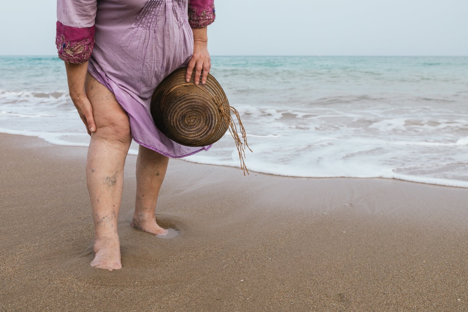 Eine Frau in einem hellvioletten Kleid steht leichtf&uuml;&szlig;ig und barfu&szlig; auf dem nassen Sand am Rande des Meeres, einen Strohhut in der Hand, w&auml;hrend sanfte Wellen anrollen - ein perfekter Sommermoment.