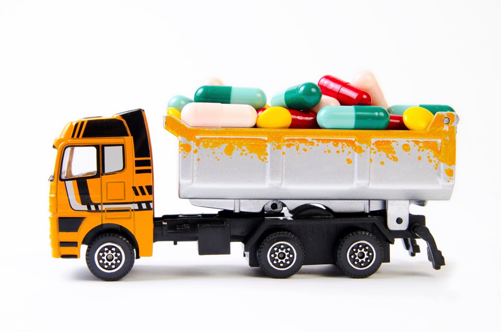 Ein Spielzeug-Muldenkipper mit orangefarbenem Fahrerhaus und silberner Ladefl&auml;che ist mit bunten Tabletten und Kapseln gef&uuml;llt, die an eine Ladung von Medikamenten oder Pillen erinnern, und steht vor einem schlichten wei&szlig;en Hintergrund.