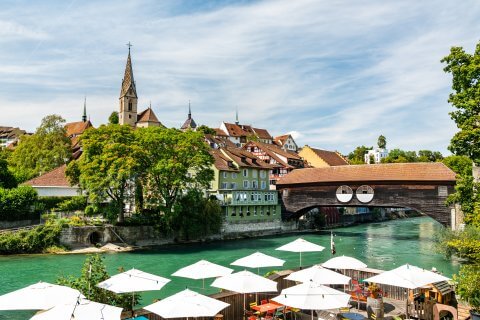 Ein malerischer Blick auf eine europ&auml;ische Stadt mit einem Kirchturm, traditionellen H&auml;usern und einer &uuml;berdachten Holzbr&uuml;cke &uuml;ber einen t&uuml;rkisfarbenen Fluss, die unter wei&szlig;en Sonnenschirmen im Au&szlig;enbereich unter einem teilweise bew&ouml;lkten Himmel 100 Jahre Badenfahrt feiert.
