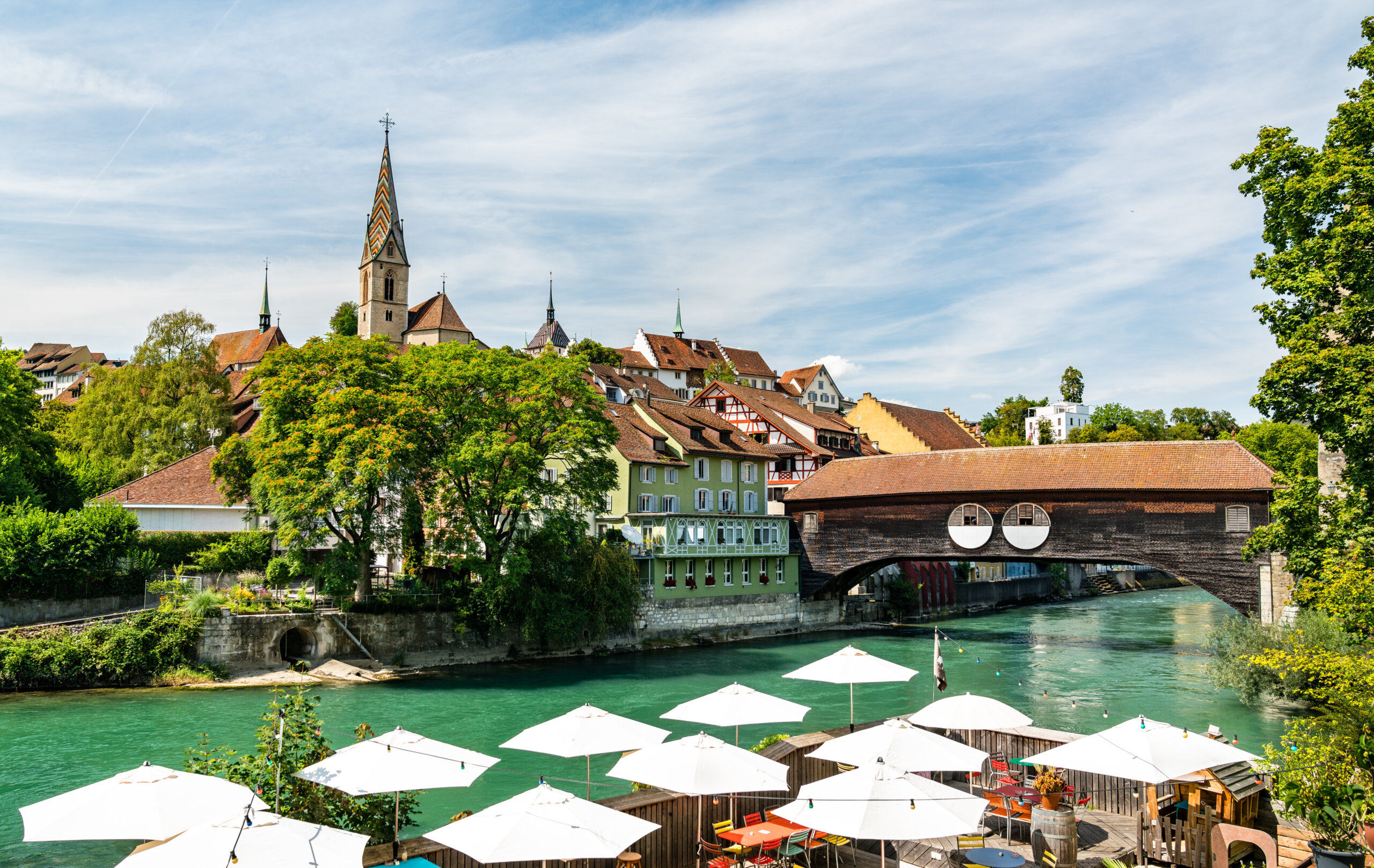 Ein malerischer Blick auf eine europ&auml;ische Stadt mit einem Kirchturm, traditionellen H&auml;usern und einer &uuml;berdachten Holzbr&uuml;cke &uuml;ber einen t&uuml;rkisfarbenen Fluss, die unter wei&szlig;en Sonnenschirmen im Au&szlig;enbereich unter einem teilweise bew&ouml;lkten Himmel 100 Jahre Badenfahrt feiert.