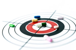 Eine Vielzahl bunter Pillen ist auf einer Zielscheibe verstreut, wobei eine gr&uuml;n-wei&szlig;e Tablette im Bullseye positioniert ist, was Pr&auml;zisionsmedizin oder gezielte Arzneimitteltherapie symbolisiert.