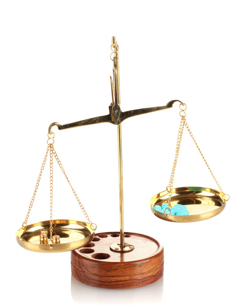 Eine Goldwaage mit Holzsockel. In der linken Schale befinden sich drei kleine Messinggewichte, in der rechten Schale zwei blaue Objekte, die m&ouml;glicherweise einer Tablette oder einem kleinen Zeh &auml;hneln. Die Waage scheint leicht geneigt zu sein, was auf eine ungleichm&auml;&szlig;ige Gewichtsverteilung hindeutet.