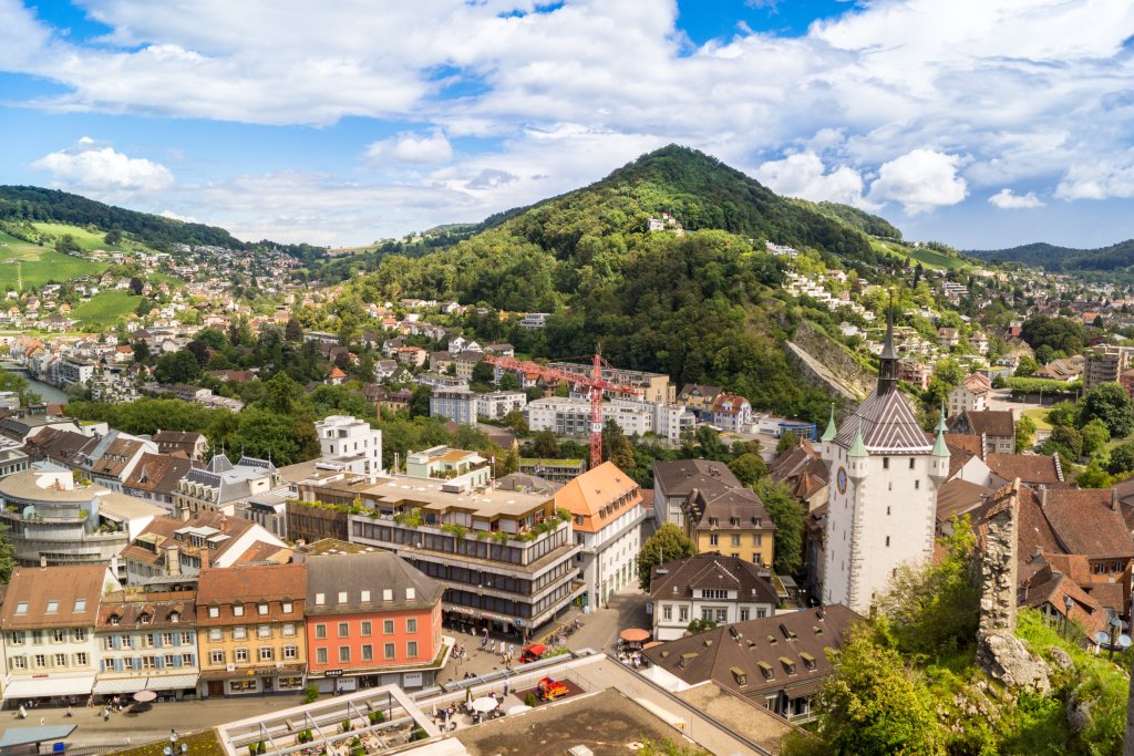 Das Panorama einer Schweizer Stadt mit farbenfrohen Geb&auml;uden, einem Kirchturm mit Uhr und &uuml;ppig gr&uuml;nen H&uuml;geln und Bergen im Hintergrund unter einem teilweise bew&ouml;lkten Himmel f&auml;ngt den Geist des 100-j&auml;hrigen Jubil&auml;ums der Badenfahrt ein.