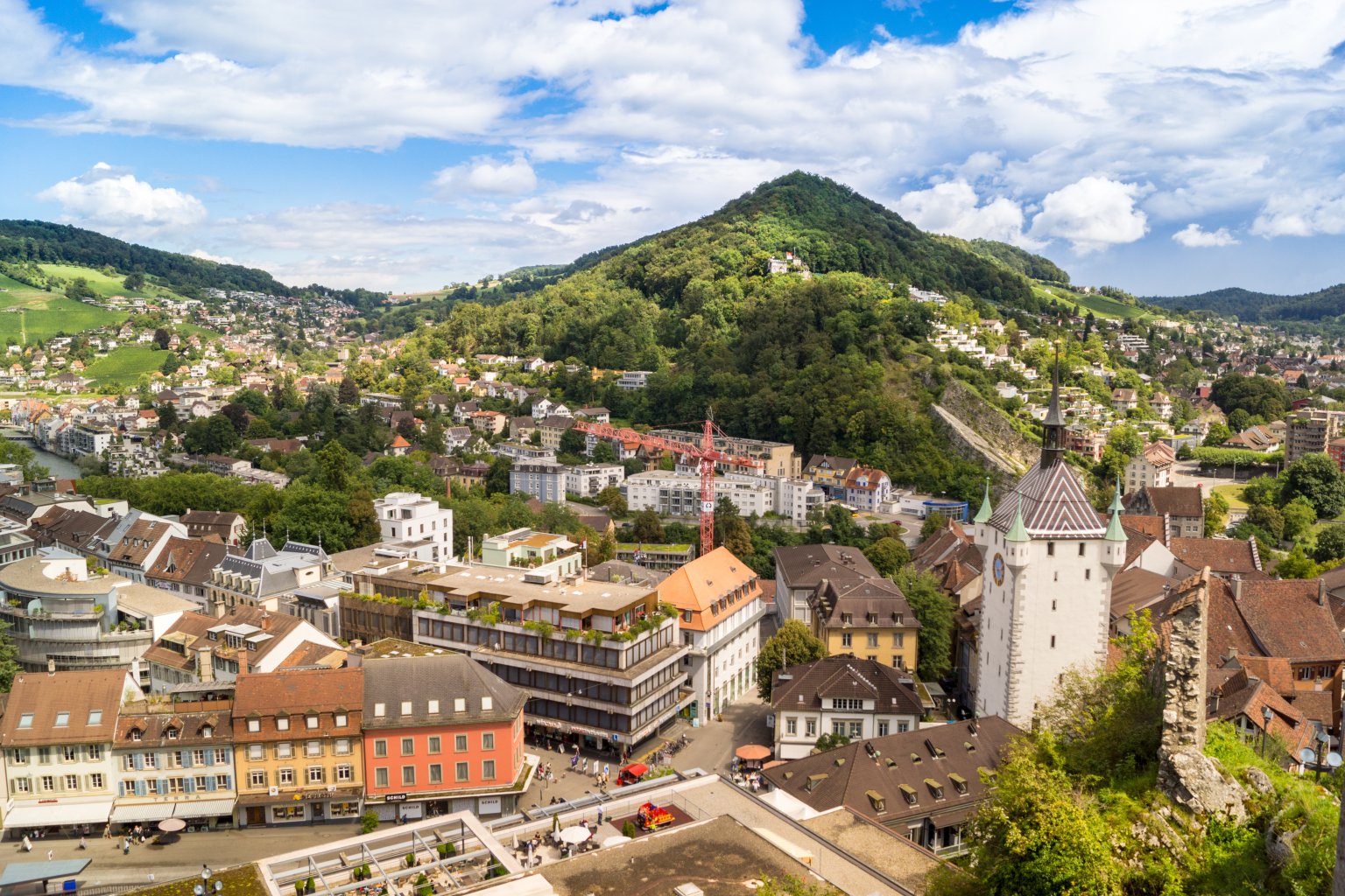 Das Panorama einer Schweizer Stadt mit farbenfrohen Geb&auml;uden, einem Kirchturm mit Uhr und &uuml;ppig gr&uuml;nen H&uuml;geln und Bergen im Hintergrund unter einem teilweise bew&ouml;lkten Himmel f&auml;ngt den Geist des 100-j&auml;hrigen Jubil&auml;ums der Badenfahrt ein.