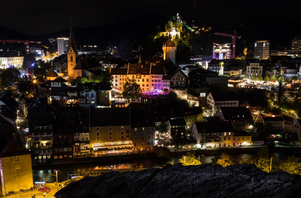 N&auml;chtliche Ansicht einer Stadt mit beleuchteten Geb&auml;uden, einschlie&szlig;lich eines Kirchturms und einer Burg auf einem H&uuml;gel. Bunte Lichter schm&uuml;cken die Stra&szlig;en anl&auml;sslich des BADENFAHRT-Jubil&auml;ums, das 100 Jahre inmitten dunkler H&uuml;gel im Hintergrund gefeiert wird.
