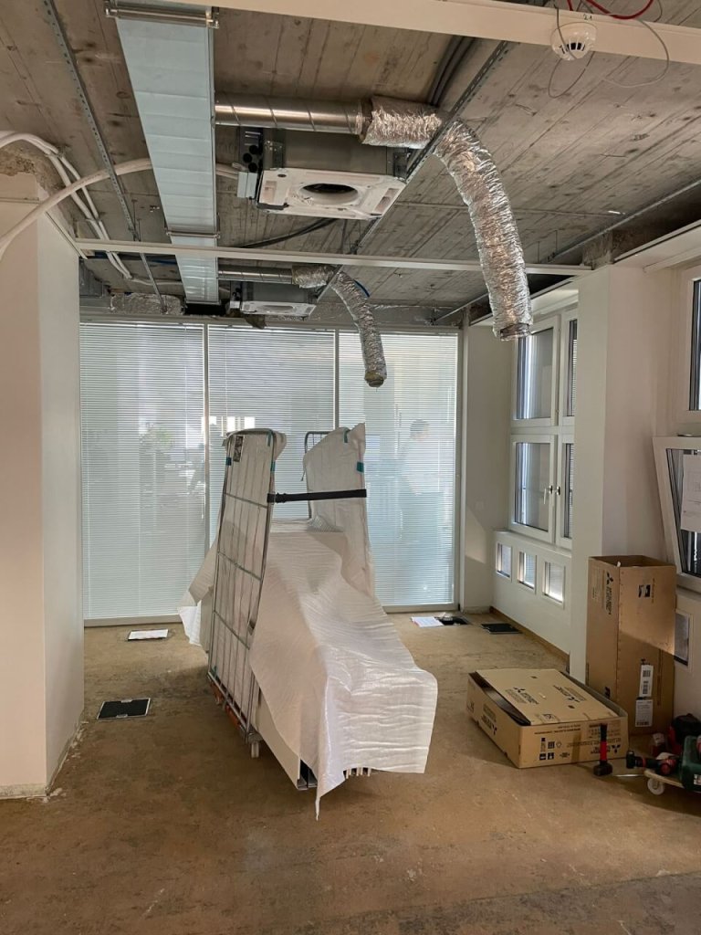 Ein unfertiger B&uuml;roraum mit freiliegenden Deckensch&auml;chten, Fensterjalousien und Kisten. Ein gro&szlig;es, mit wei&szlig;em Kunststoff &uuml;berzogenes Objekt steht in der Mitte, und Baumaterialien - Schl&uuml;ssel f&uuml;r den QMS kontinuierlichen Verbesserungsprozess - sind im Raum verstreut.