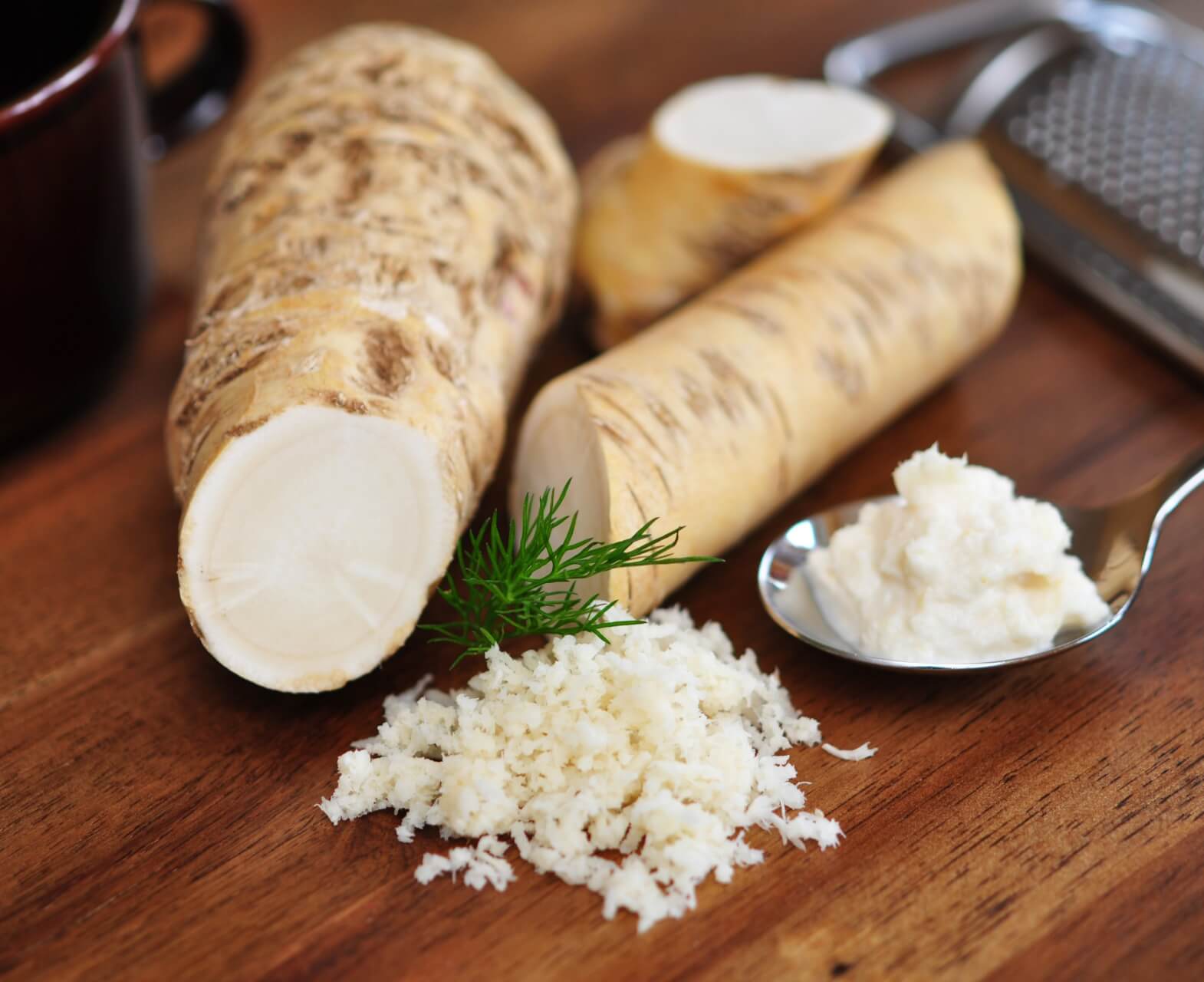 Ein Holztisch mit ganzen Meerrettichwurzeln, ein Haufen frisch geriebener Meerrettich, ein L&ouml;ffel cremiger So&szlig;e, ein Dillzweig und eine Metallreibe im Hintergrund.