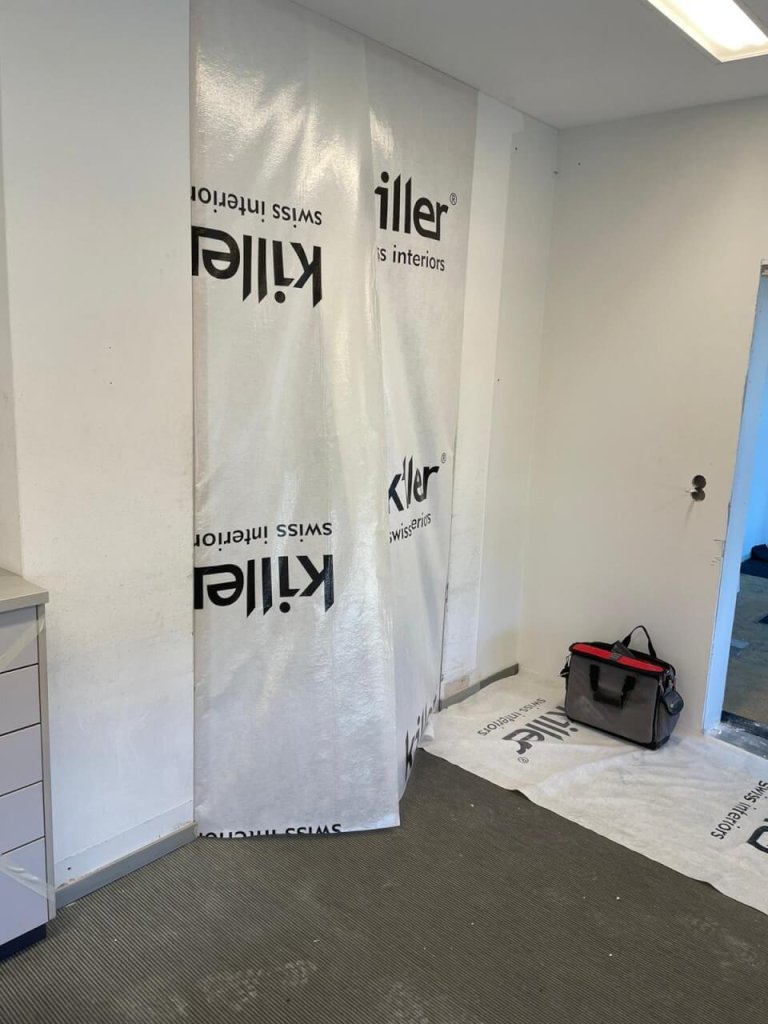 Ein wei&szlig;er Raum, der gerade renoviert wird, mit einer gro&szlig;en Plastikfolie mit der Aufschrift Killer SWISS interiors an einer Wand, spiegelt Kundenorientierung wider. Eine Werkzeugtasche liegt auf einer Folie auf dem Boden neben einer offenen T&uuml;r und verdeutlicht einen kontinuierlichen Verbesserungsprozess.