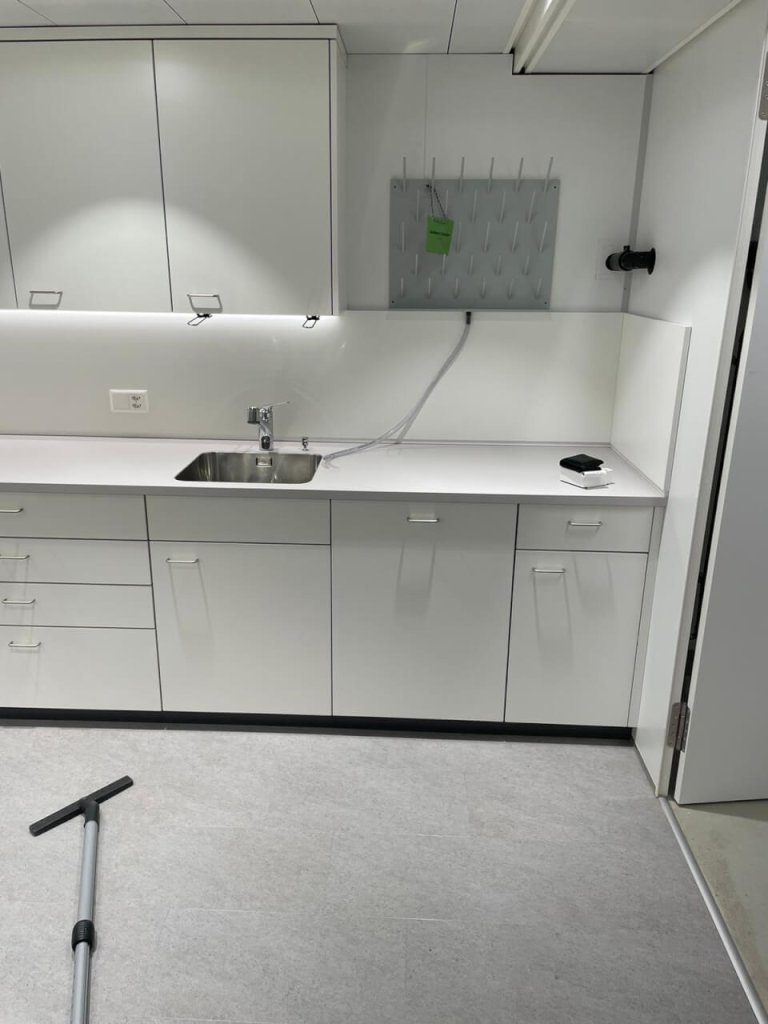 Ein sauberer, moderner wei&szlig;er Labor- oder Medizinraum mit Schr&auml;nken, einem Waschbecken und einem auf QMS ausgerichteten Layout. Eine an der Wand befestigte Stecktafel und ein Wischmoppgriff auf dem hellgrauen Boden spiegeln Kundenorientierung und einen kontinuierlichen Verbesserungsprozess wider. Der Raum ist aufger&auml;umt und gut beleuchtet.