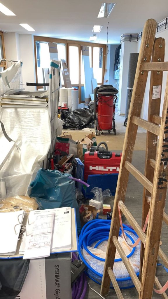 Ein unordentlicher Raum, der gerade renoviert wird, mit Werkzeugen, einer Leiter, Baumaterialien, blauen Kabeln, einem roten Werkzeugkasten, Papieren, Taschen und verstreuten Ger&auml;ten spiegelt den kontinuierlichen Verbesserungsprozess wider, wobei nat&uuml;rliches Licht durch die Fenster im Hintergrund f&auml;llt.