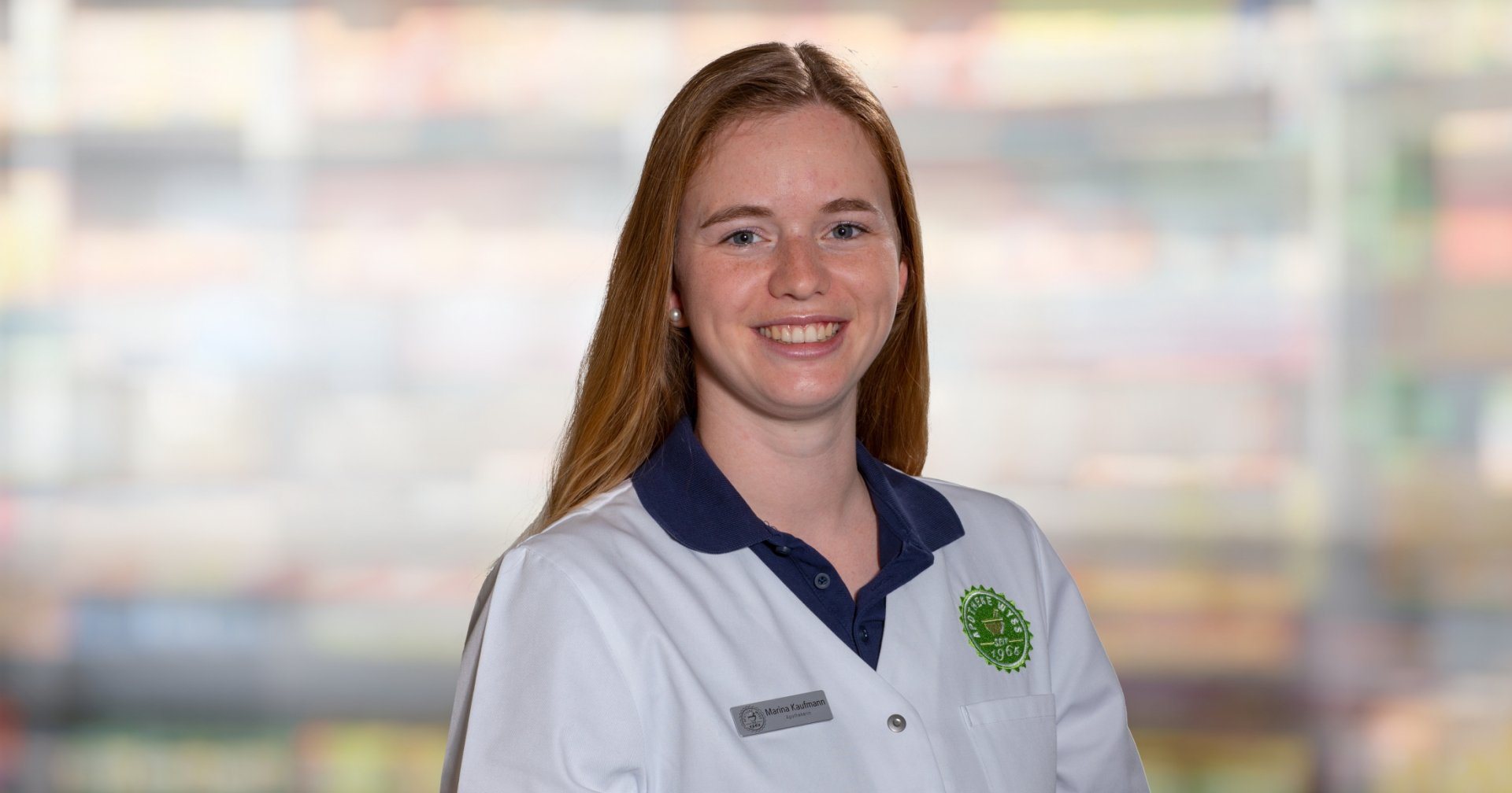 Eine Frau mit langen roten Haaren l&auml;chelt, w&auml;hrend sie einen wei&szlig;en Laborkittel mit einem gr&uuml;nen Logo und einem Namensschild mit der Aufschrift "Marina Kaufmann" tr&auml;gt. Im verschwommenen Hintergrund sind bunte, unscharfe Regale zu sehen.