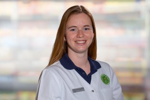 Eine Frau mit langen roten Haaren l&auml;chelt, w&auml;hrend sie einen wei&szlig;en Laborkittel mit einem gr&uuml;nen Logo und einem Namensschild mit der Aufschrift "Marina Kaufmann" tr&auml;gt. Im verschwommenen Hintergrund sind bunte, unscharfe Regale zu sehen.