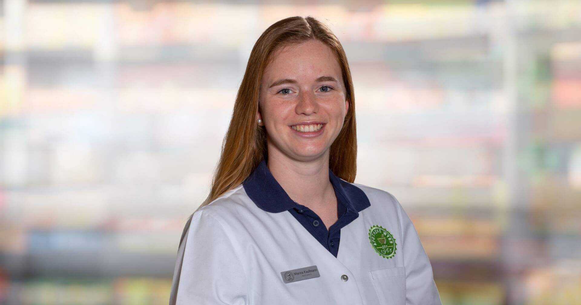 Eine Frau mit langen roten Haaren l&auml;chelt, w&auml;hrend sie einen wei&szlig;en Laborkittel mit einem gr&uuml;nen Logo und einem Namensschild mit der Aufschrift "Marina Kaufmann" tr&auml;gt. Im verschwommenen Hintergrund sind bunte, unscharfe Regale zu sehen.