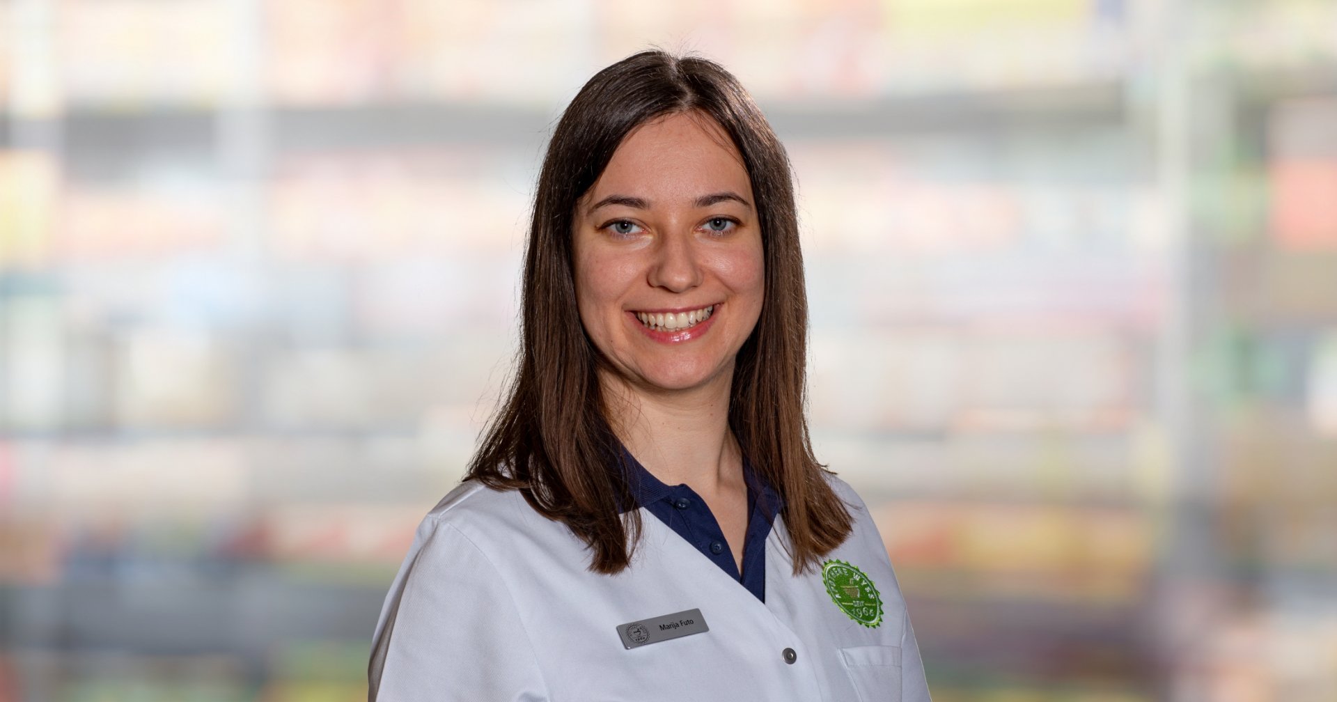 Eine Frau mit glattem braunem Haar, die einen wei&szlig;en Laborkittel und ein Namensschild tr&auml;gt, l&auml;chelt in die Kamera. Der Hintergrund ist unscharf und zeigt bunte Regale, die auf einen Laden oder eine Apotheke hindeuten.