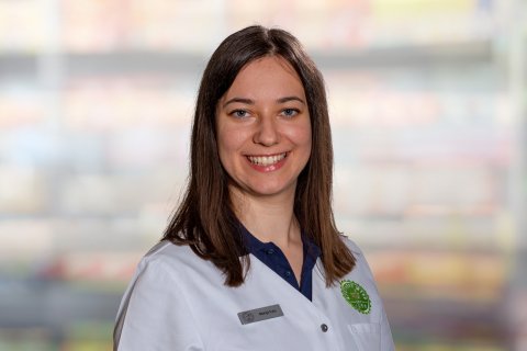 Eine Frau mit glattem braunem Haar, die einen wei&szlig;en Laborkittel und ein Namensschild tr&auml;gt, l&auml;chelt in die Kamera. Der Hintergrund ist unscharf und zeigt bunte Regale, die auf einen Laden oder eine Apotheke hindeuten.