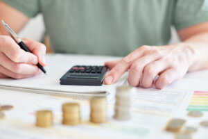 Eine Person benutzt einen Taschenrechner, w&auml;hrend sie in ein Notizbuch schreibt, mit Stapeln von M&uuml;nzen und Finanzdokumenten auf dem Tisch, was an Budgetierung oder Finanzplanung denken l&auml;sst, etwa um bei den Krankenkassen-Pr&auml;mien 2024 zu sparen.