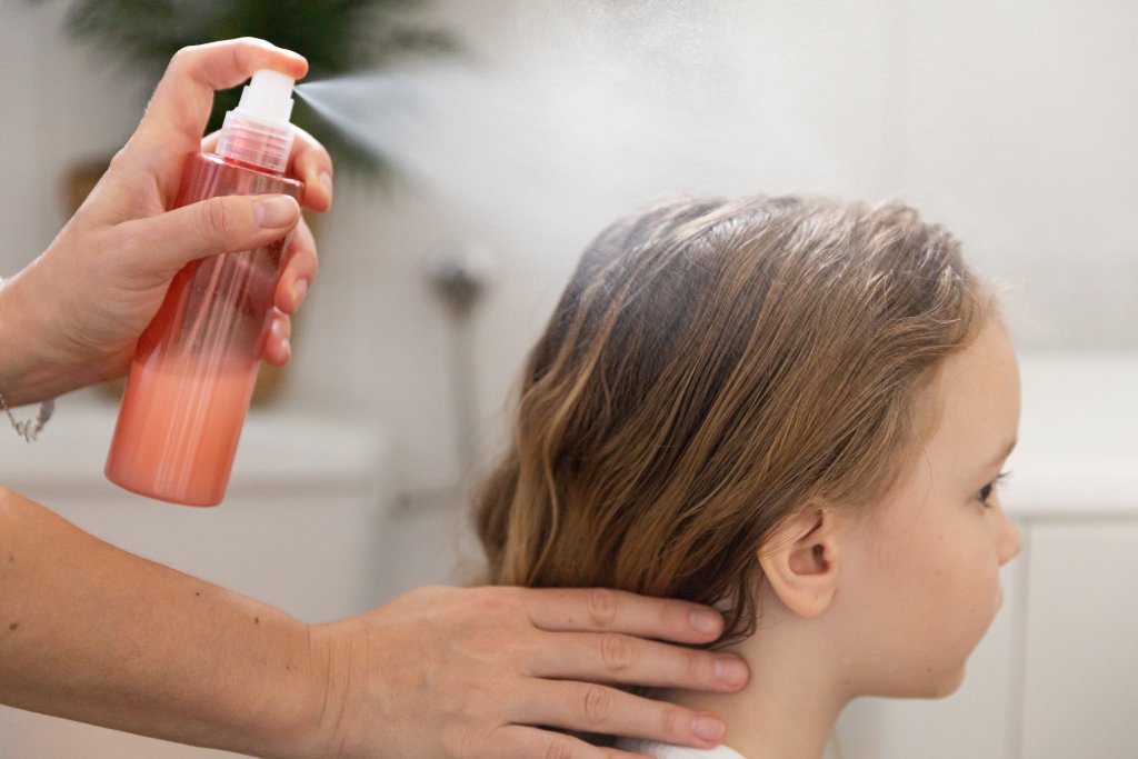 Eine Person spr&uuml;ht Leave-in-Conditioner aus einer Flasche auf das Haar eines kleinen Kindes mit hellbraunem Haar und h&auml;lt dabei sanft den Nacken des Kindes, um die Herausforderung von Kopfl&auml;use und das Haar gesund zu halten zu meistern.