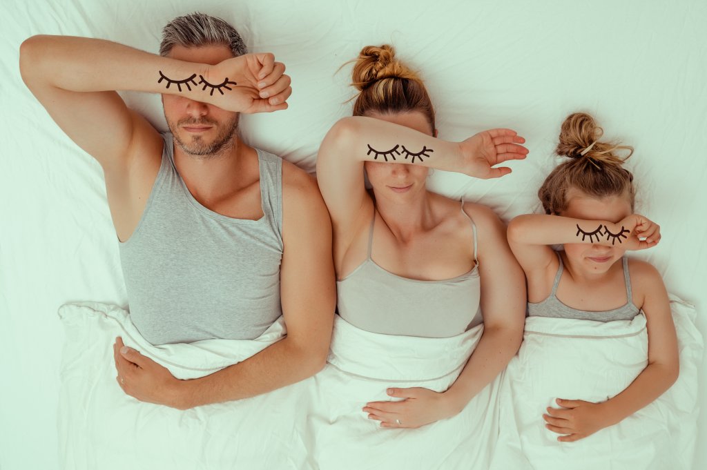 Ein Mann, eine Frau und ein Kind liegen in einem Bett mit wei&szlig;en Laken und bedecken ihre Augen mit jeweils einem Arm. Wimpernillustrationen auf ihren Armen imitieren geschlossene Augen und unterstreichen die Bedeutung des k&ouml;rpereigenen Schlafs als Teil unseres Regenerationssystems.