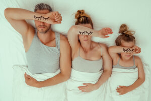 Ein Mann, eine Frau und ein Kind liegen in einem Bett mit wei&szlig;en Laken und bedecken ihre Augen mit jeweils einem Arm. Wimpernillustrationen auf ihren Armen imitieren geschlossene Augen und unterstreichen die Bedeutung des k&ouml;rpereigenen Schlafs als Teil unseres Regenerationssystems.