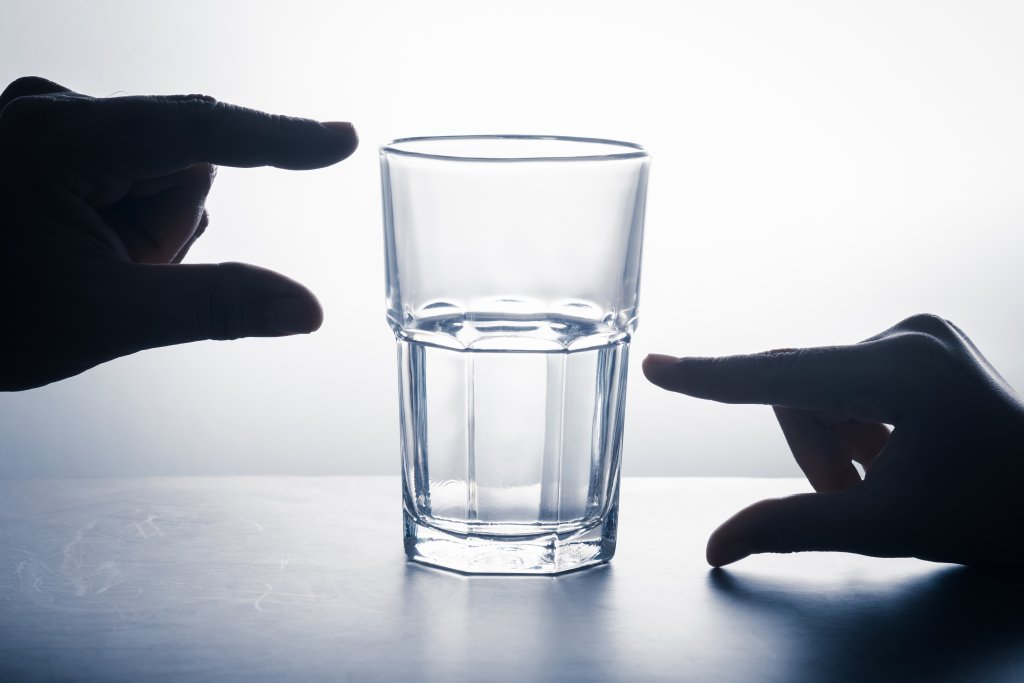 Ein Glas Wasser steht auf einer Fl&auml;che, eine Hand liegt auf jeder Seite des Glases. Der Hintergrund ist hell und die H&auml;nde und das Glas sind als Silhouette dargestellt, was Gesundheit und die Macht der Gedanken im t&auml;glichen Leben symbolisiert.