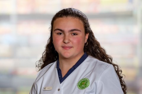 Eine junge Person mit langen lockigen Haaren tr&auml;gt eine wei&szlig;e Uniform mit einem gr&uuml;nen Emblem und einem Namensschild und steht vor einem unscharfen Hintergrund.