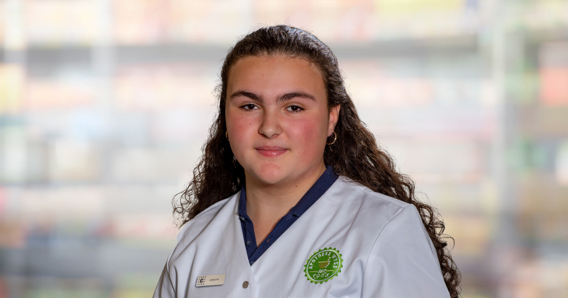 Eine junge Person mit langen lockigen Haaren tr&auml;gt eine wei&szlig;e Uniform mit einem gr&uuml;nen Emblem und einem Namensschild und steht vor einem unscharfen Hintergrund.