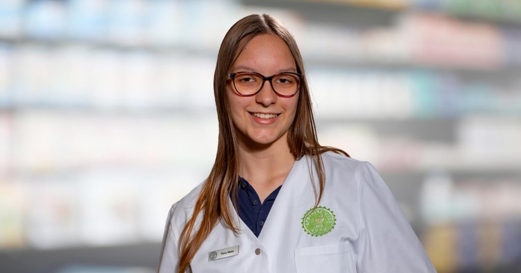 Eine junge Frau mit langen braunen Haaren und Brille l&auml;chelt, w&auml;hrend sie einen wei&szlig;en Apothekenkittel und ein Namensschild tr&auml;gt und vor verschwommenen, mit Produkten gef&uuml;llten Regalen steht.