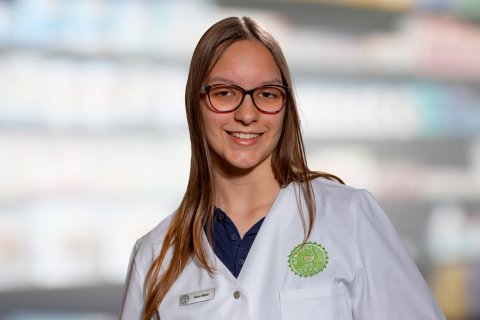 Eine junge Frau mit langen braunen Haaren und Brille l&auml;chelt, w&auml;hrend sie einen wei&szlig;en Apothekenkittel und ein Namensschild tr&auml;gt und vor verschwommenen, mit Produkten gef&uuml;llten Regalen steht.
