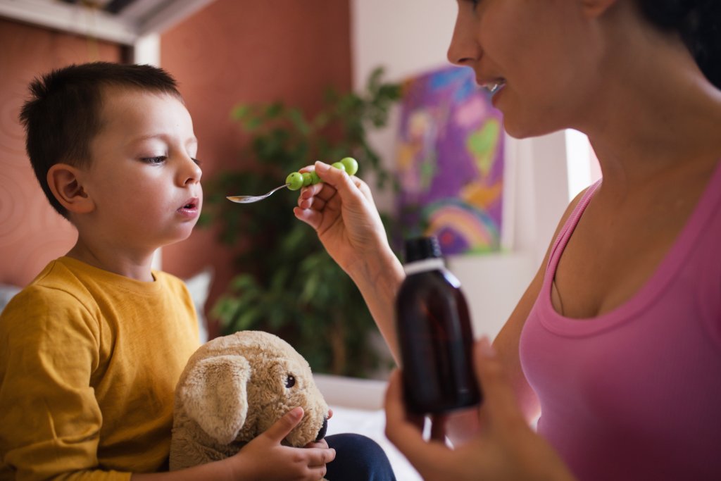 Eine Frau in einem rosafarbenen Oberteil gibt einem kleinen Jungen, der ein Stofftier in der Hand h&auml;lt, Medizin - ihr spezielles Elixier. Der Junge in einem gelben Hemd sitzt drinnen und schaut auf den L&ouml;ffel, w&auml;hrend ein buntes Gem&auml;lde und eine gr&uuml;ne Pflanze den Hintergrund erhellen.