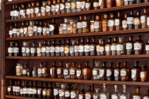 Die Regale sind gef&uuml;llt mit alten Apothekerflaschen aus Glas in verschiedenen Gr&ouml;&szlig;en, beschriftet mit medizinischen Namen und geheimnisvollen Elixieren. Die Flaschen enthalten Fl&uuml;ssigkeiten und Pulver in bernsteinfarbenen, braunen und klaren T&ouml;nen.