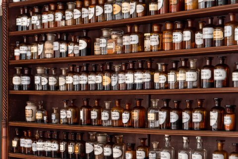 Die Regale sind gef&uuml;llt mit alten Apothekerflaschen aus Glas in verschiedenen Gr&ouml;&szlig;en, beschriftet mit medizinischen Namen und geheimnisvollen Elixieren. Die Flaschen enthalten Fl&uuml;ssigkeiten und Pulver in bernsteinfarbenen, braunen und klaren T&ouml;nen.