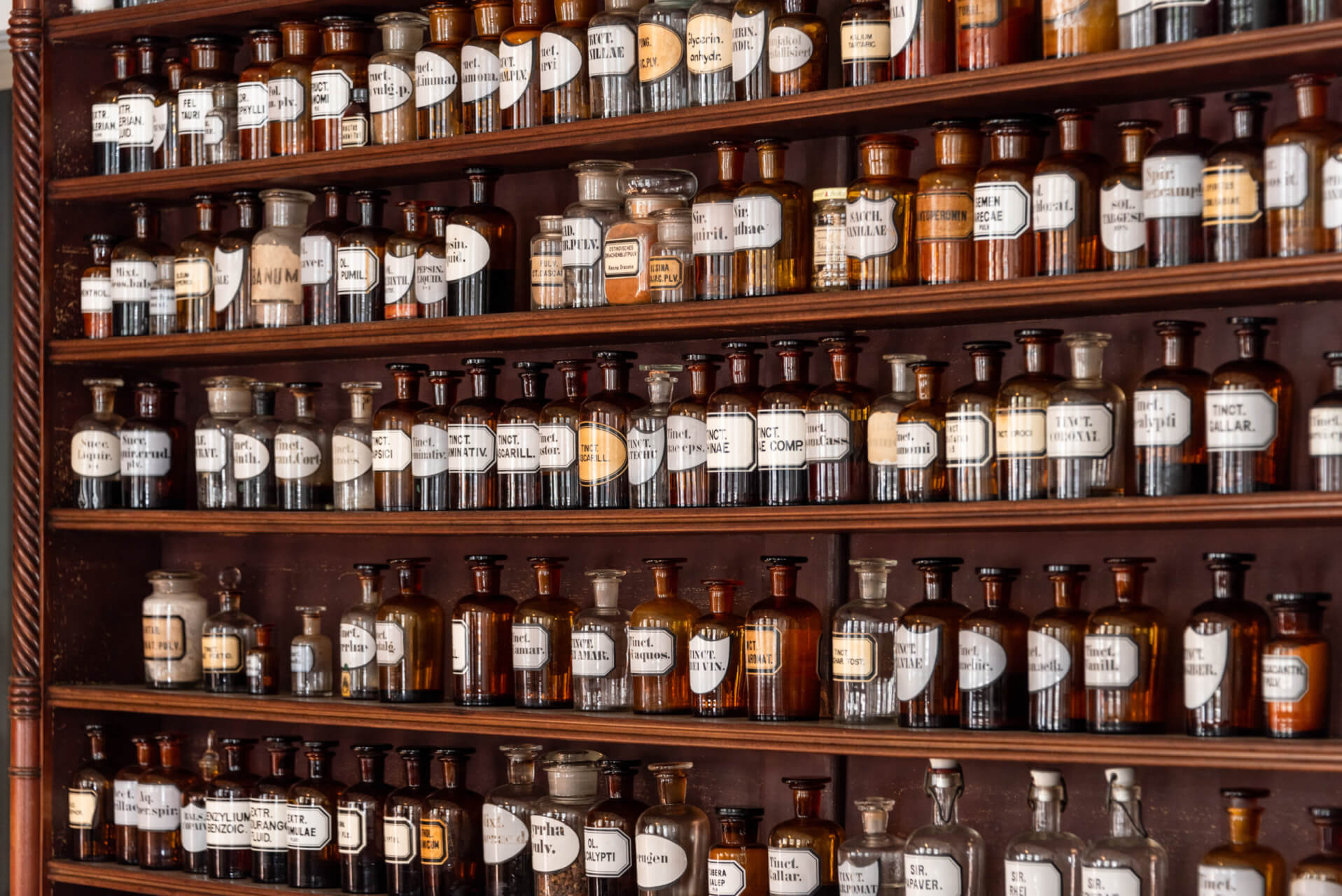 Die Regale sind gef&uuml;llt mit alten Apothekerflaschen aus Glas in verschiedenen Gr&ouml;&szlig;en, beschriftet mit medizinischen Namen und geheimnisvollen Elixieren. Die Flaschen enthalten Fl&uuml;ssigkeiten und Pulver in bernsteinfarbenen, braunen und klaren T&ouml;nen.