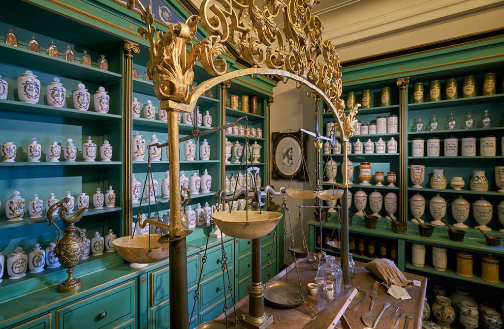 Eine alte Apotheke mit tealfarbenen Regalen, die mit verschn&ouml;rkelten Gl&auml;sern ausgekleidet sind, einer goldenen Waage auf einem h&ouml;lzernen Tresen und verschiedenen antiken Glasflaschen und Elixieren, die fein s&auml;uberlich im Vordergrund stehen.