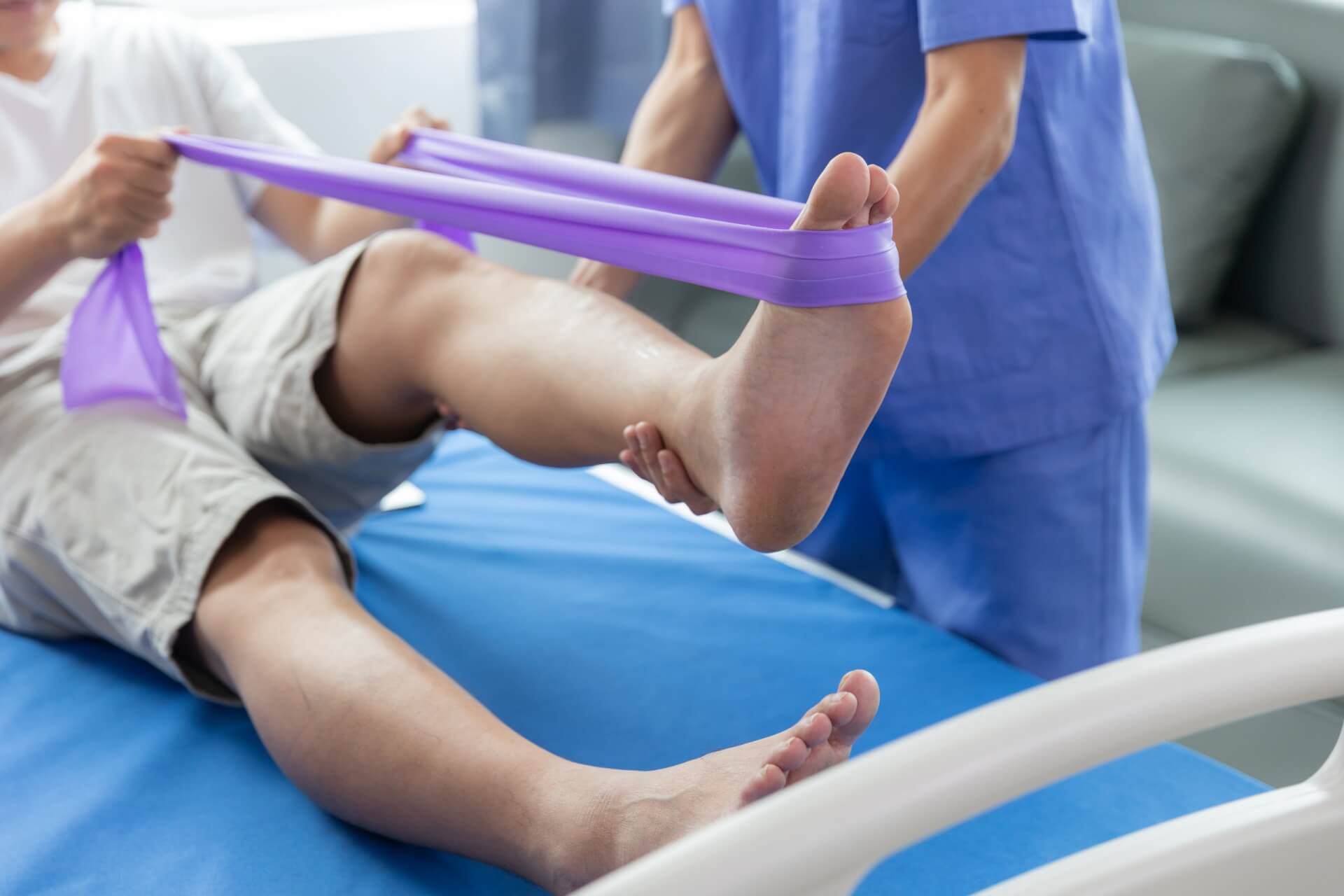 Eine Person in kurzen Hosen f&uuml;hrt eine Bein&uuml;bung mit einem lila Widerstandsband durch, unterst&uuml;tzt von einer medizinischen Fachkraft in blauer Kleidung auf einem Krankenbett - ein effektiver Ansatz zur Behandlung von Gelenkbeschwerden in einer klinischen Umgebung.