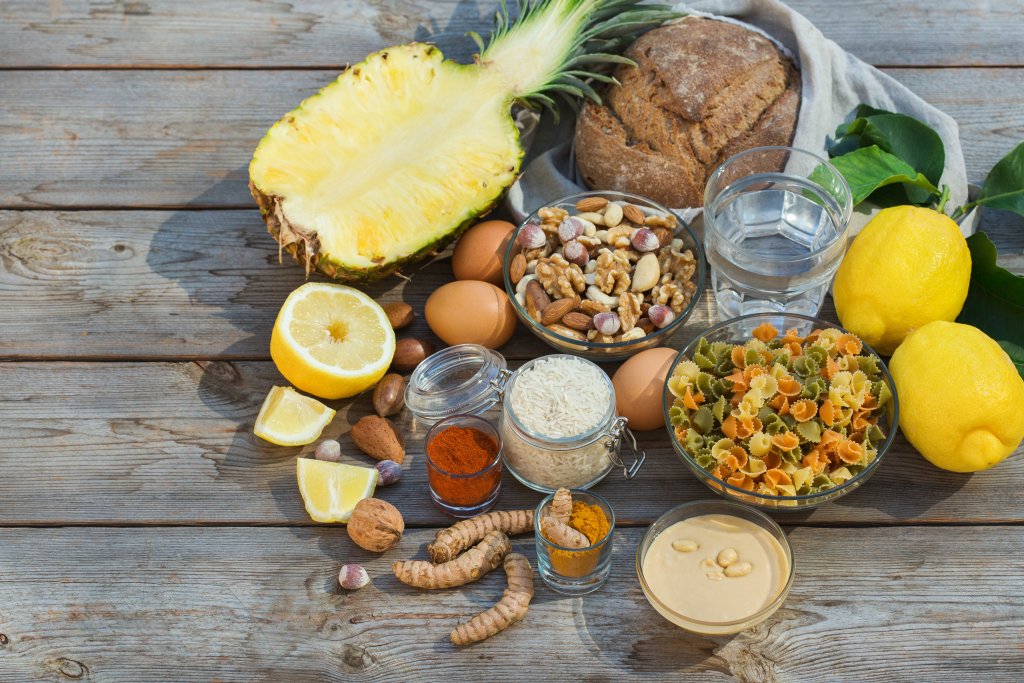 Eine Vielzahl von Lebensmitteln auf einem Holztisch, darunter eine halbe Ananas, Zitronen, Brot, Eier, gemischte N&uuml;sse, Reis, Nudeln, Gl&auml;ser mit Wasser, Gl&auml;ser mit Gew&uuml;rzen, Kurkumawurzeln und eine Schale mit Tahini.