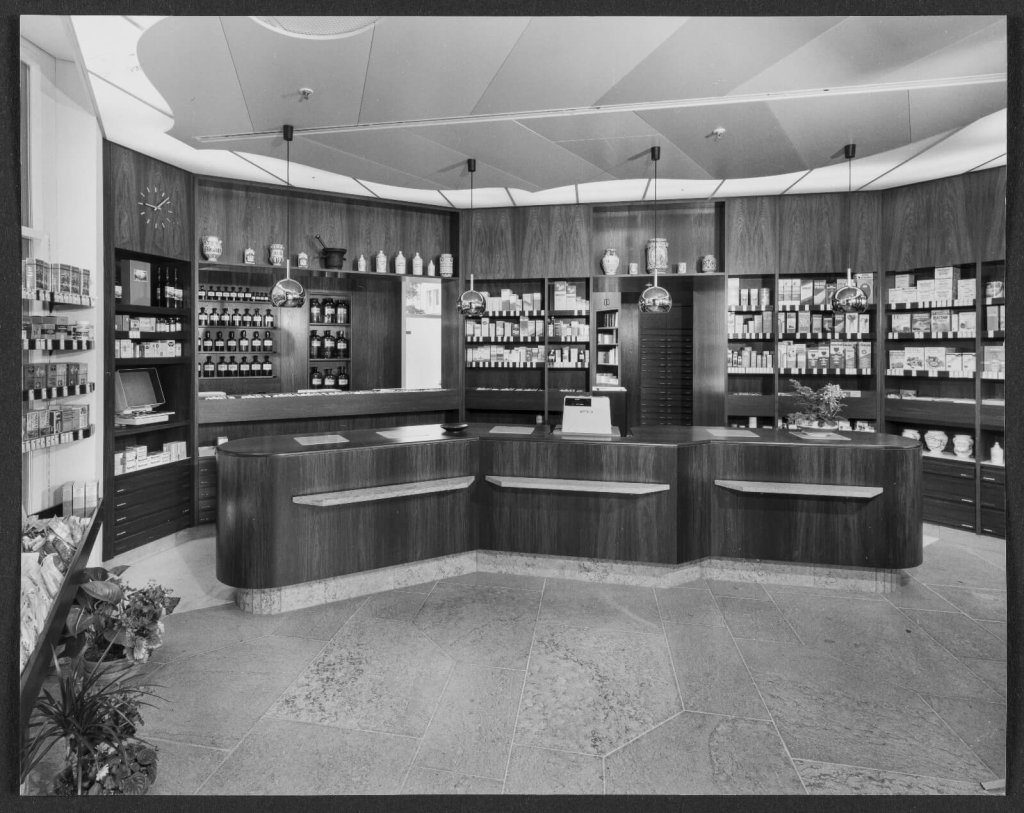 Schwarz-Weiss-Foto des Innenraums der alten Apotheke Wyss in der N&auml;he des Bahnhofs Baden, mit Holzregalen, die mit Flaschen, Kisten und Gl&auml;sern gef&uuml;llt sind. In der Mitte steht ein gro&szlig;er Tresen mit einer Registrierkasse, w&auml;hrend H&auml;ngelampen den Raum beleuchten.