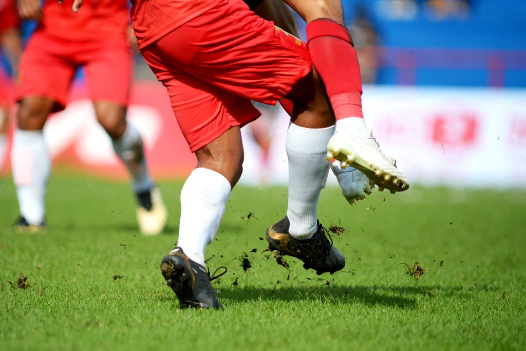 Nahaufnahme von zwei Fu&szlig;ballspielern in roten Shorts und wei&szlig;en Stutzen, die auf einer Wiese um den Ball k&auml;mpfen, wobei Dreck aufgewirbelt wird, als der eine den anderen angreift - was sowohl den Nervenkitzel als auch das Risiko von Sportverletzungen ohne entsprechende Pr&auml;ventionstipps verdeutlicht.