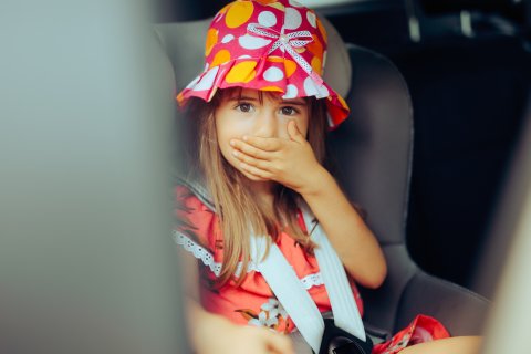 Ein junges M&auml;dchen mit farbenfrohem Hut und rotem Kleid sitzt auf einem Autositz, h&auml;lt sich den Mund mit der Hand zu und schaut mit gro&szlig;en Augen nach vorne - vielleicht sp&uuml;rt es die ersten Anzeichen einer Reisekrankheit.