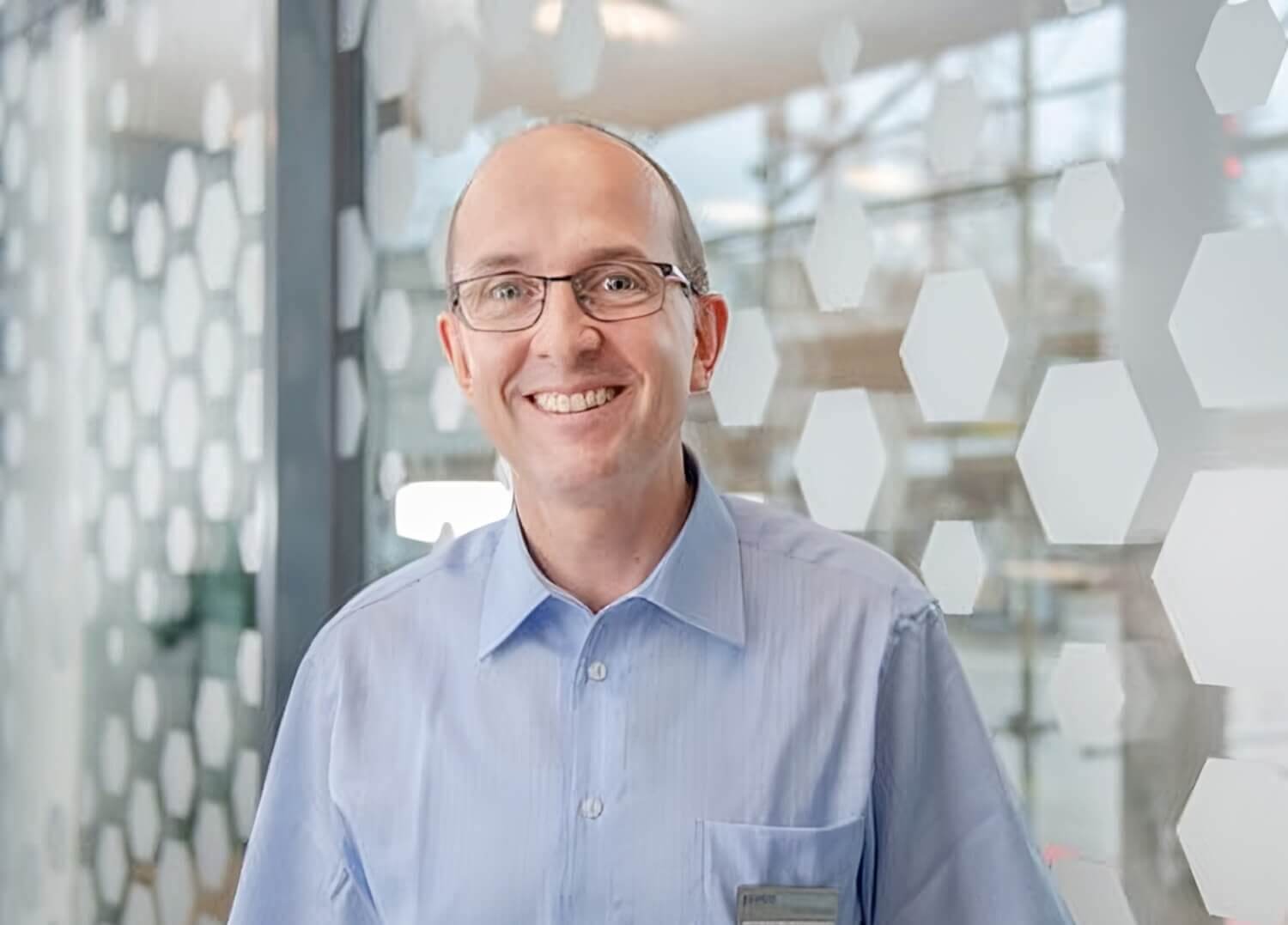 Ein l&auml;chelnder Mann, Dr. med. Martin Geyer, der eine Brille und ein hellblaues Hemd mit Kragen tr&auml;gt, steht in einem Innenraum vor einer Glaswand mit wei&szlig;en sechseckigen Mustern, bereit f&uuml;r ein Interview &uuml;ber Darmgesundheit.