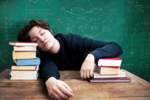 Eine schlafende Person, deren Kopf auf einem Stapel B&uuml;cher an einem Schreibtisch ruht, mit Mathe- und Wissenschaftsformeln auf einer Kreidetafel im Hintergrund.