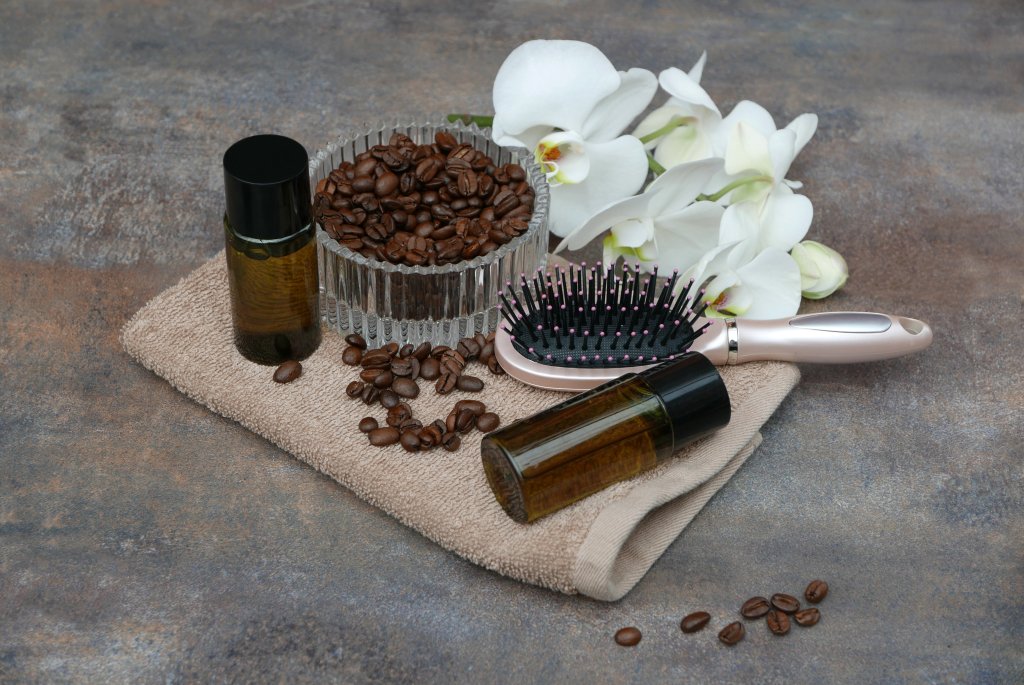 Eine Glasschale mit Kaffeebohnen, zwei kleine Flaschen, eine Haarb&uuml;rste und wei&szlig;e Orchideen sind auf einem beigen Handtuch vor einem grauen Hintergrund angeordnet.