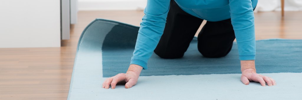Ein &auml;lterer Erwachsener in blauem Hemd und dunkler Hose liegt auf H&auml;nden und Knien auf einem blauen Teppich und scheint sich vom Boden zu erheben, um die Bedeutung der Sturzpr&auml;vention am Unfallort Zuhause zu verdeutlichen. Nur der Oberk&ouml;rper und die Arme sind sichtbar.
