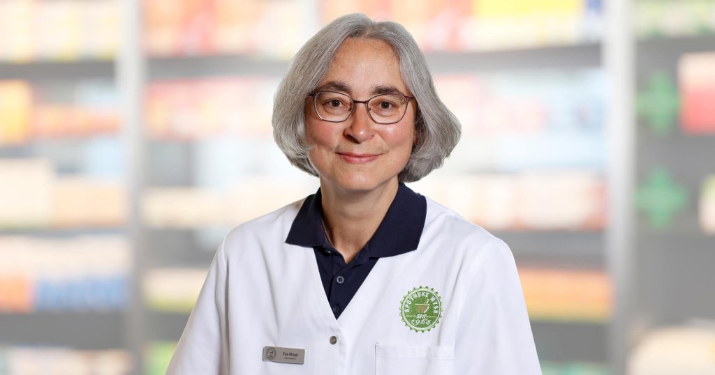 Ein Apotheker mit grauem Haar und Brille, der einen wei&szlig;en Laborkittel mit gr&uuml;nem Apothekenlogo tr&auml;gt, steht in einem Ladengang mit unscharfen Regalen mit Produkten im Hintergrund.